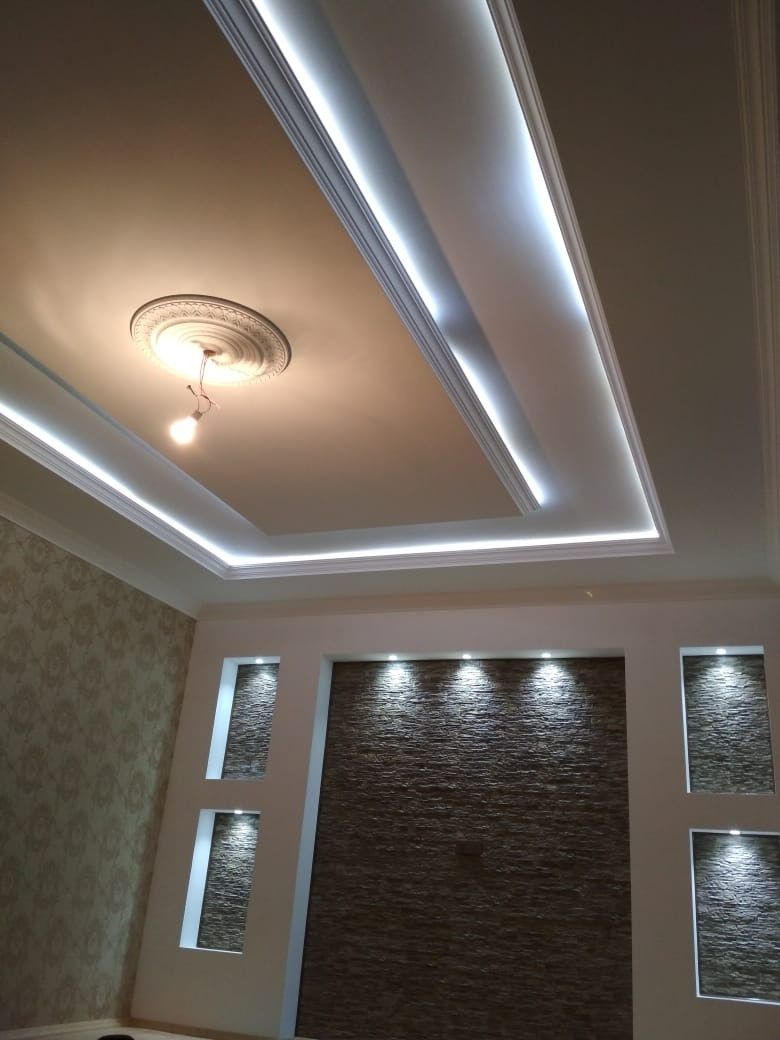 Nowoczesny sufit wielopoziomowy z oświetleniem LED, ozdobny element centralny z żarówką, ściana z dekoracyjną okładziną imitującą kamień z wnękami podświetlonymi punktowo.