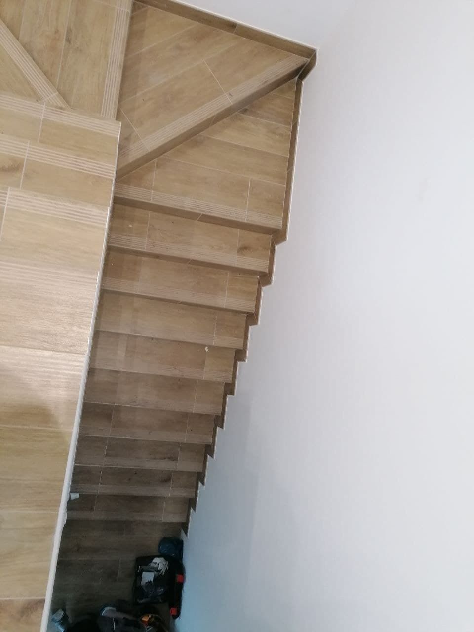 Schody wyłożone płytkami ceramicznymi imitującymi drewno, widok z góry, przy białej ścianie, z widocznymi narzędziami i workami na dole.