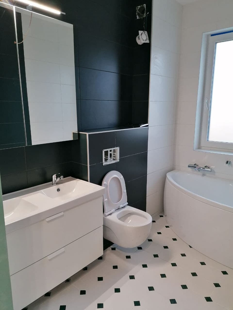 Wykończona łazienka w czarno-białej kolorystyce z wanną narożną, wiszącą toaletą z otwartą klapą i szafką z umywalką, widoczne czarne płytki na ścianach i białe płytki podłogowe z czarnymi...