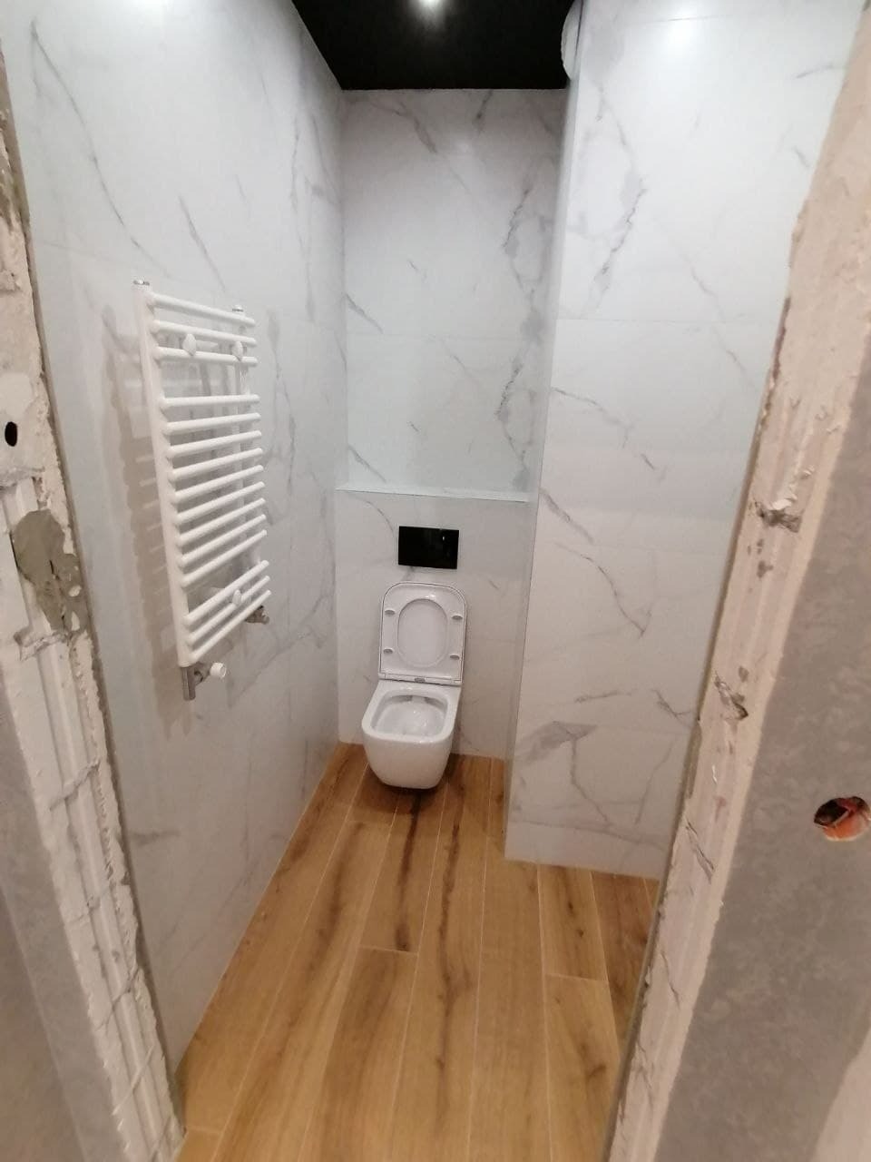 Wąska łazienka po remoncie z białymi marmurkowymi płytkami, drewnopodobną podłogą, wiszącą toaletą i grzejnikiem drabinkowym, widok z niedokończonego wejścia.