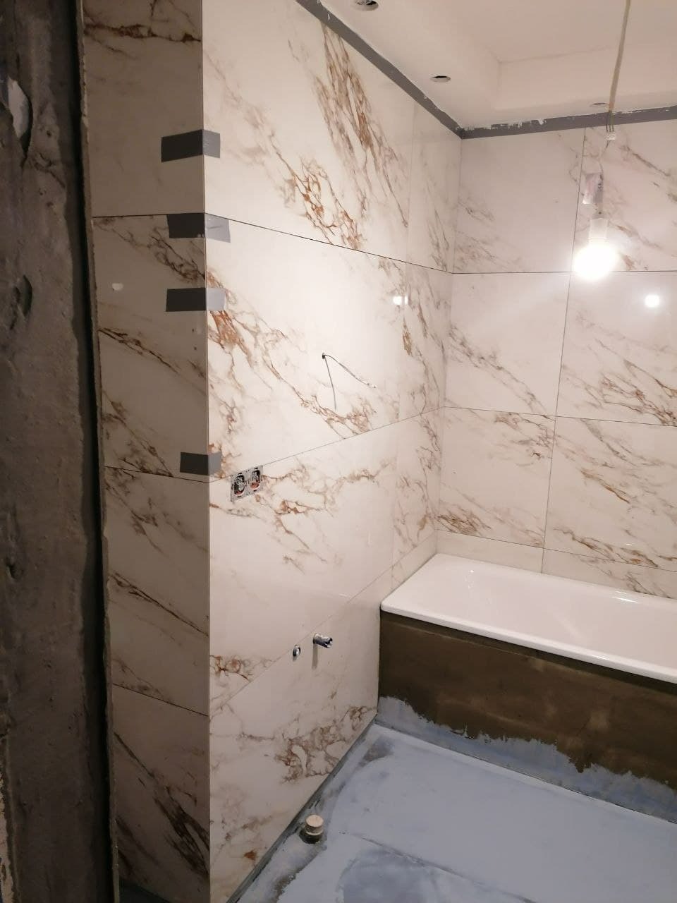 Płytki imitujące marmur na ścianach łazienki w trakcie montażu, zabezpieczone taśmą, z widoczną białą wanną i przygotowanym podłożem pod posadzkę.