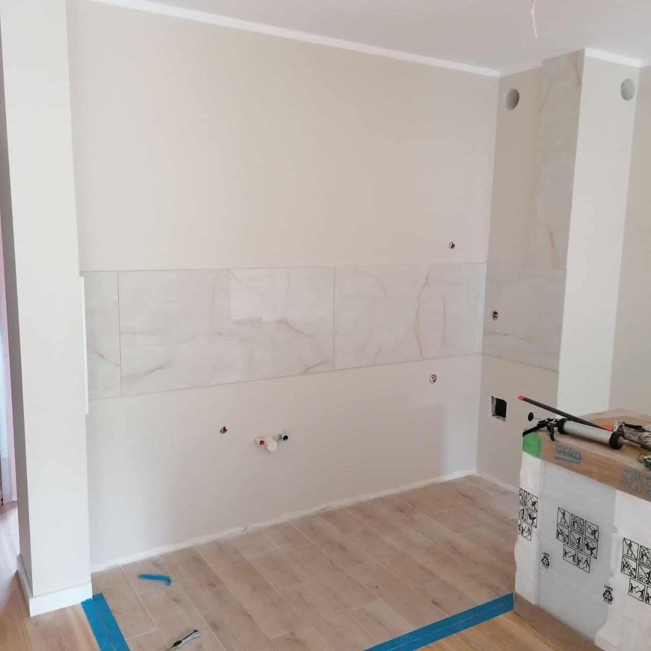 Wnętrze kuchni w trakcie wykańczania: położone płytki imitujące marmur na ścianie, widoczne otwory na instalacje wodno-kanalizacyjne, podłoga wyłożona panelami drewnopodobnymi, zabezpieczona...