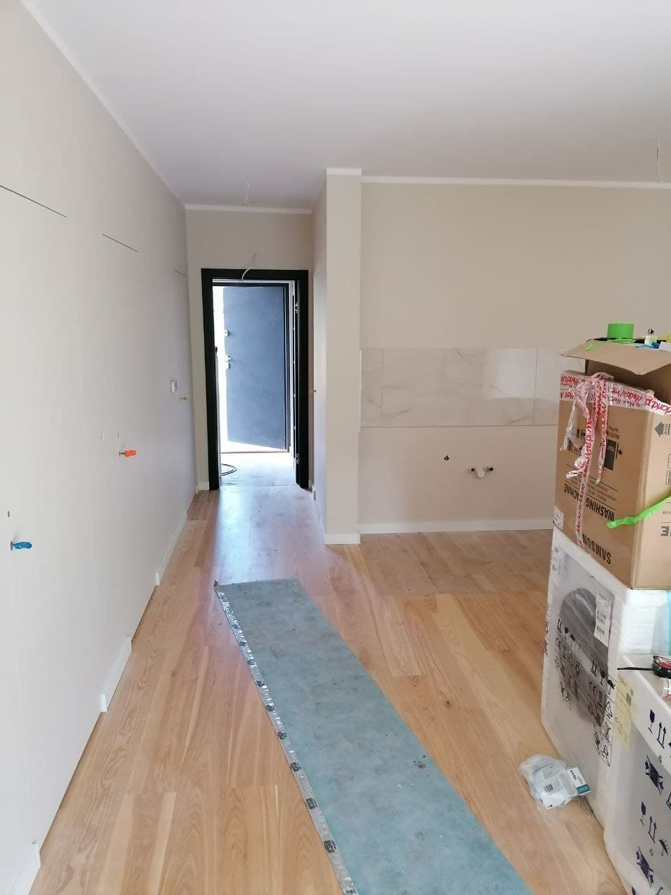 Wnętrze mieszkania w trakcie remontu: świeżo położona drewniana podłoga, białe ściany z widocznymi przewodami, zabezpieczona folią podłoga, kartony ze sprzętem AGD, widok na otwarte drzwi.