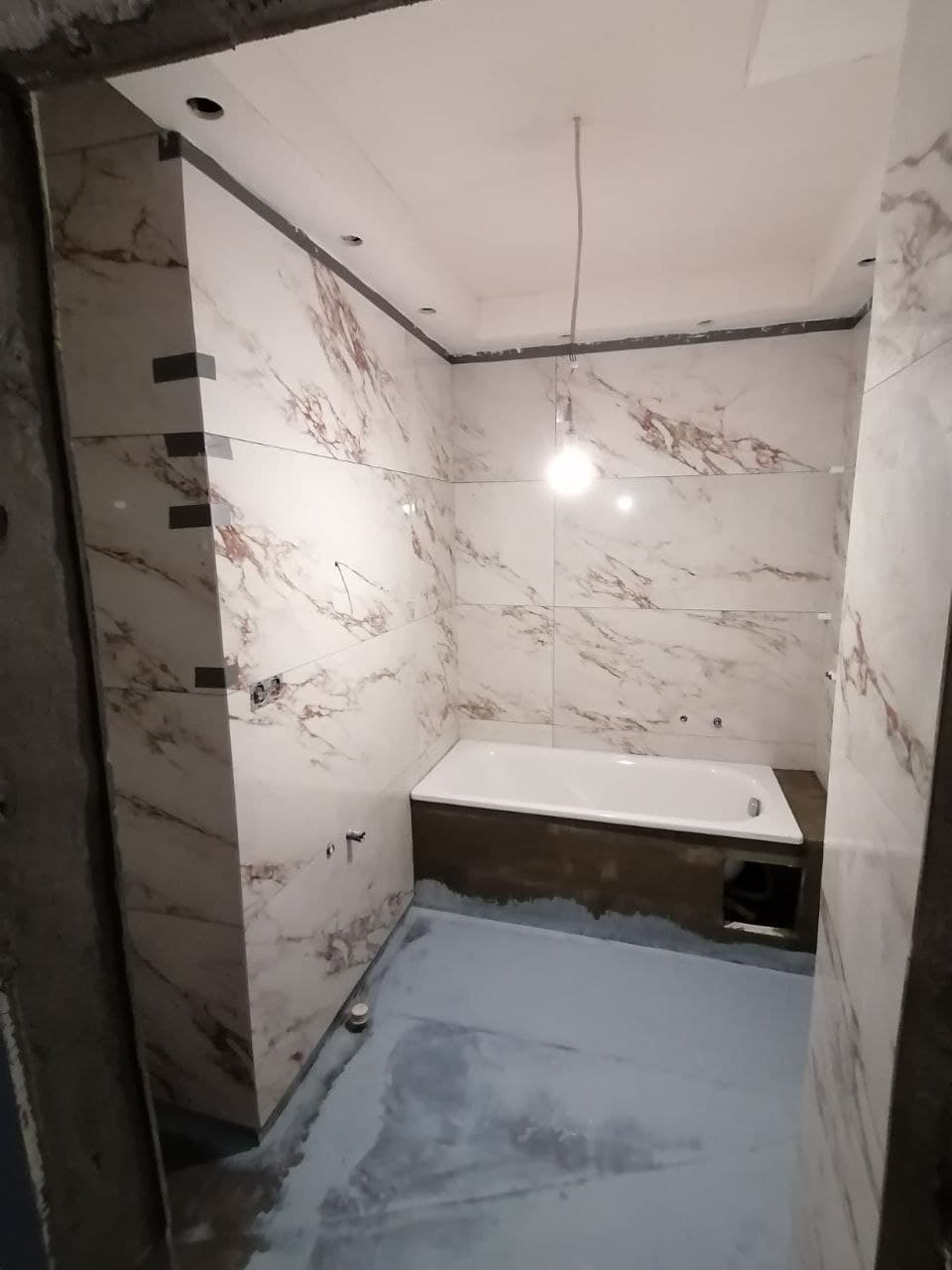 Wnętrze łazienki w trakcie remontu: biała wanna obudowana ciemnym drewnem, ściany wyłożone marmurowymi płytkami, podłoga pokryta szarą wylewką, widoczne niedokończone prace i przewód z żarówką...