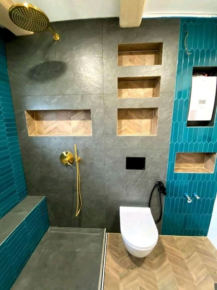 Wykończona łazienka z szarymi płytkami imitującymi kamień, złotą armaturą prysznicową, turkusowymi płytkami heksagonalnymi, drewnopodobną podłogą w jodełkę i wnękami w ścianach.