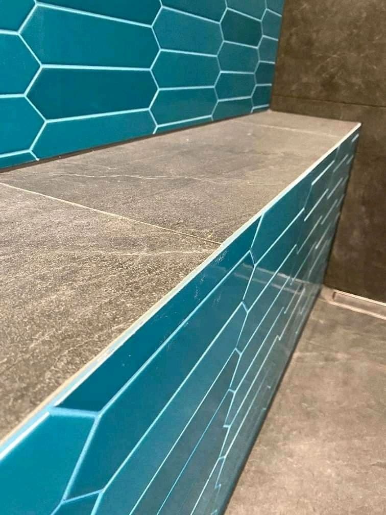 Wykończona półka w łazience wyłożona turkusowymi, heksagonalnymi płytkami z białymi fugami, przylegająca do szarej, kamiennej powierzchni, z widocznym odpływem liniowym w podłodze.