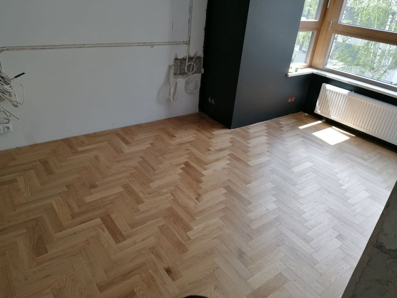 Ukończona podłoga z parkietu ułożonego w jodełkę w pokoju z oknem, grzejnikiem i przygotowanymi przewodami elektrycznymi na ścianie.