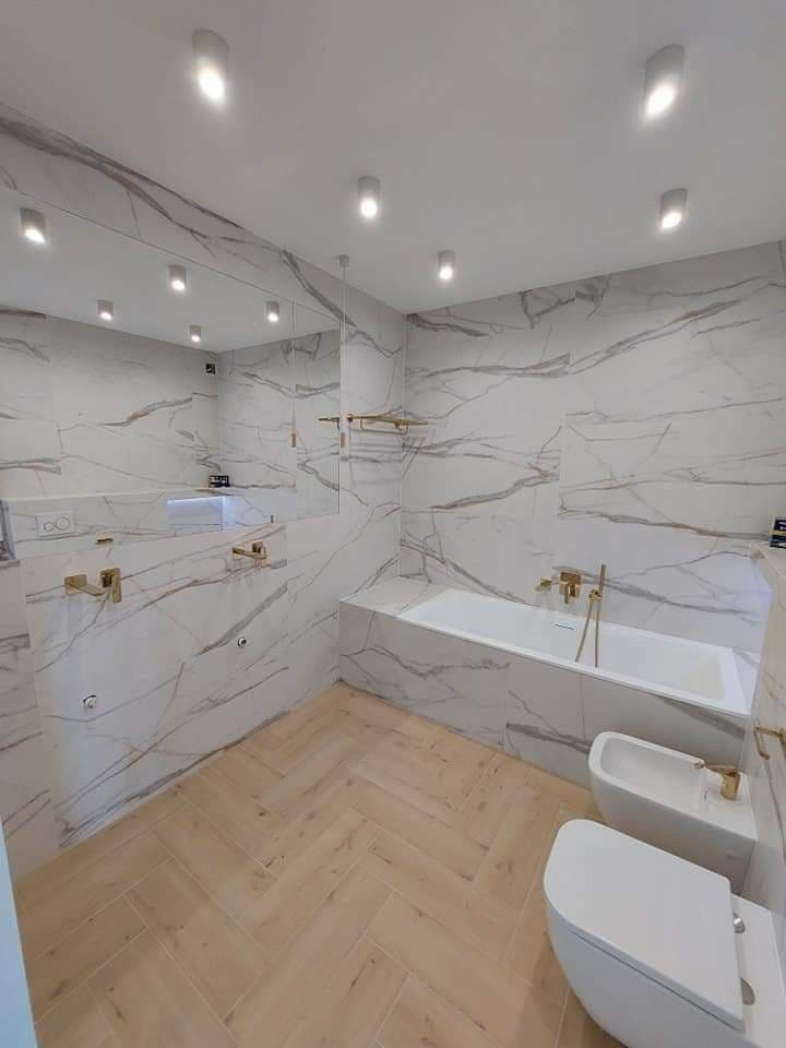 Elegancka łazienka z białymi płytkami imitującymi marmur z szarymi żyłami, drewnopodobną podłogą ułożoną w jodełkę, złotą armaturą i oświetleniem punktowym.