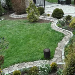 Ujęcie z góry na zadbany ogród z wijącą się ścieżką z kamieni, obrzeżami z kostki, trawnikiem, ozdobnym pawiem i dekoracyjnym pniem drzewa