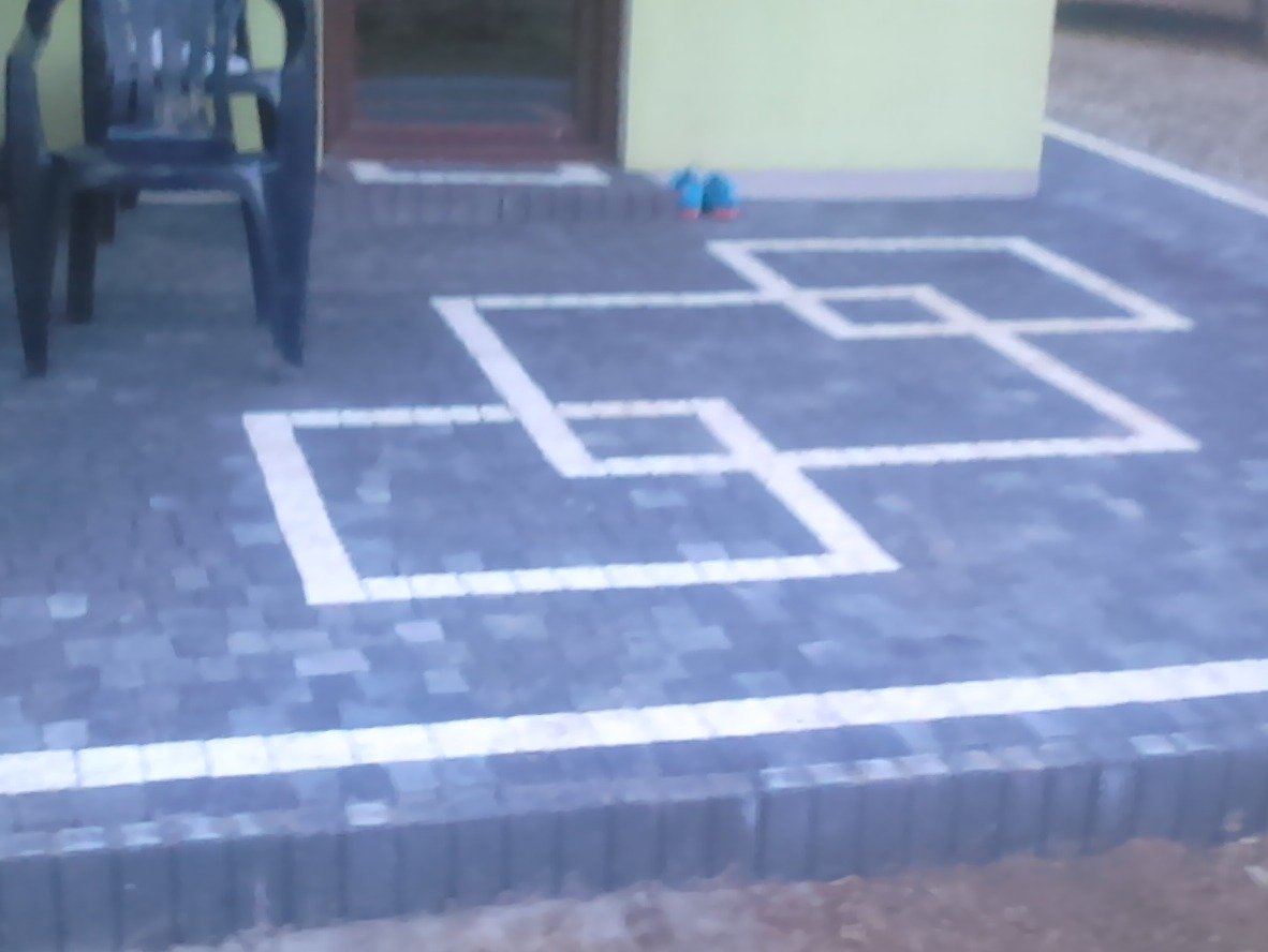 Ułożona szara kostka brukowa z białym wzorem geometrycznym kwadratów przed wejściem do domu z żółtą elewacją, widoczny czarny plastikowy fotel i buty przed drzwiami.