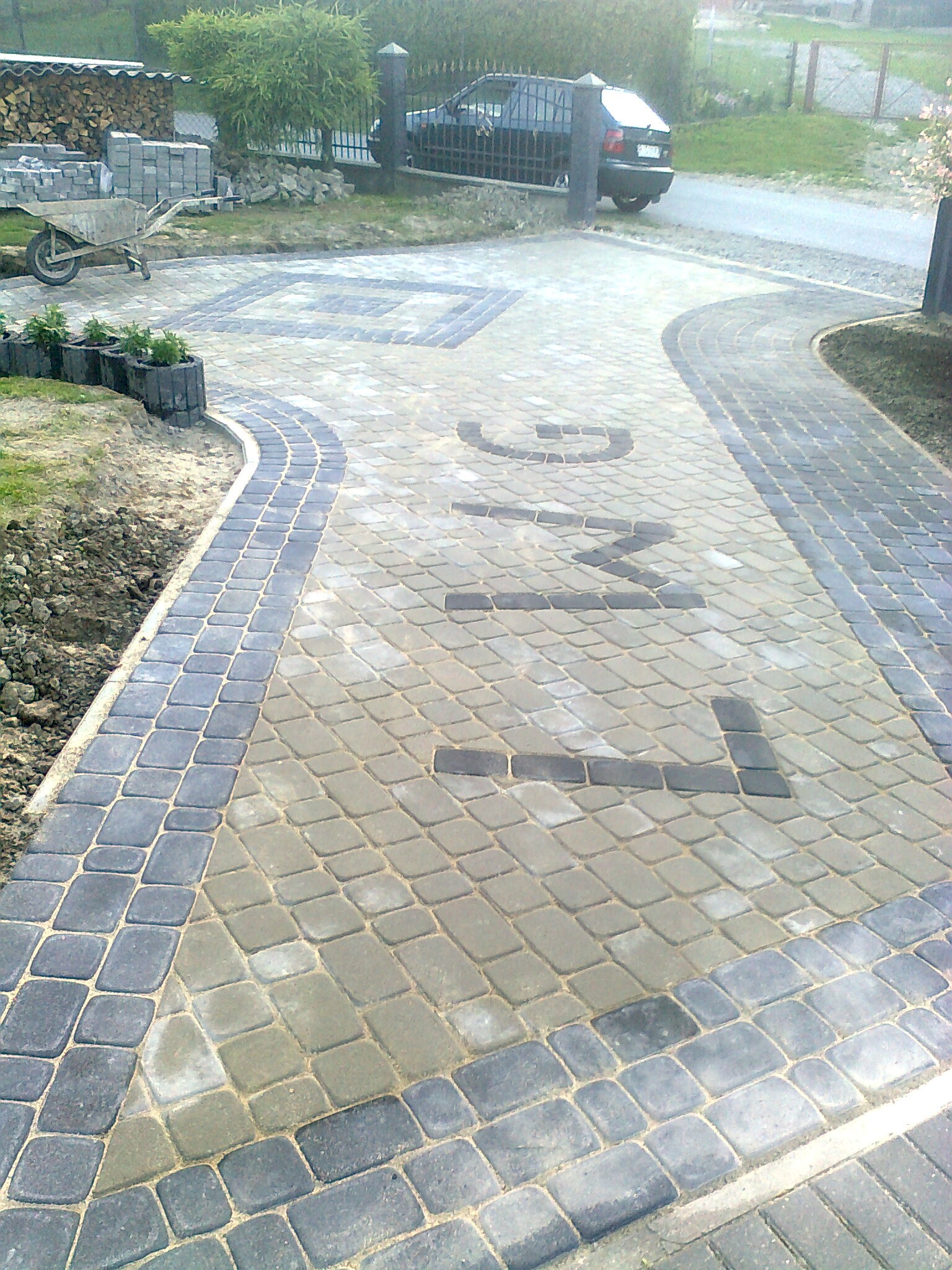 Ułożony z kostki brukowej podjazd z napisem 'KING' wkomponowanym w nawierzchnię, obrzeża z ciemnej kostki, w tle zaparkowany samochód i składowane materiały budowlane.