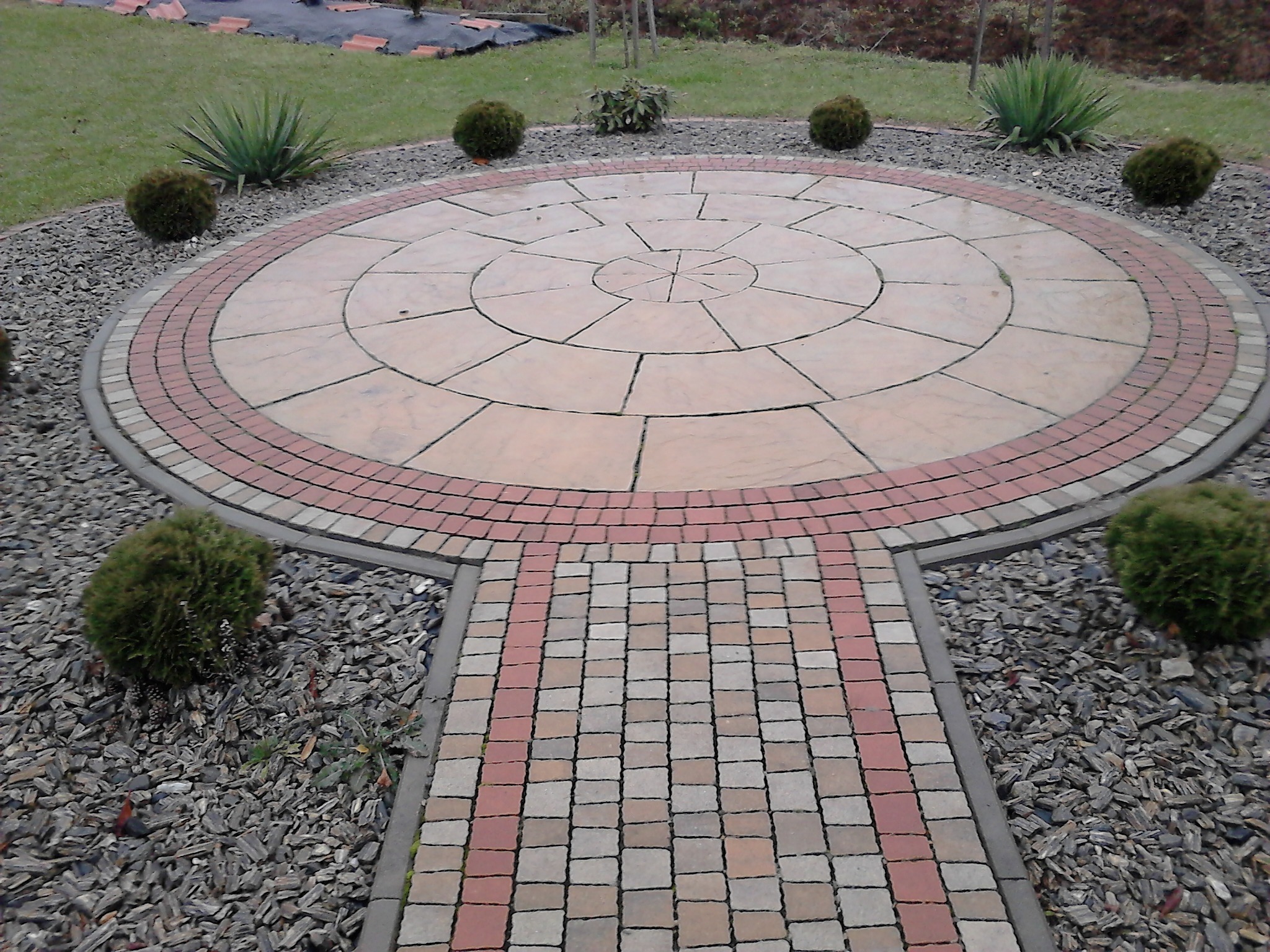 Okrągły plac z kostki brukowej w Dobryni, otoczony ozdobnym żwirem i kulistymi krzewami, z wybrukowanym chodnikiem prowadzącym do niego.