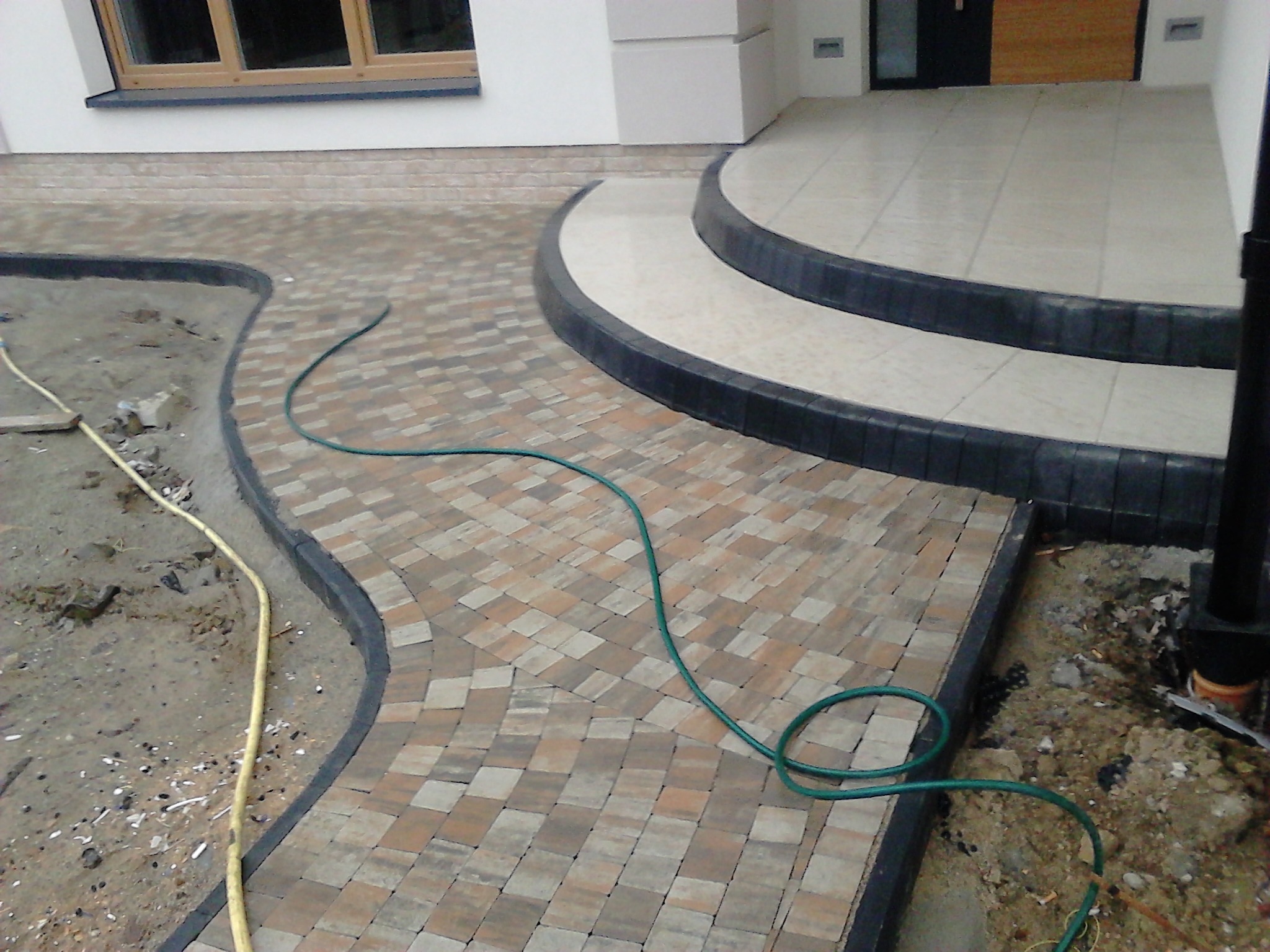 Ułożona kostka brukowa w odcieniach beżu i brązu prowadząca do schodów wejściowych domu, widoczne węże ogrodowe na ziemi.
