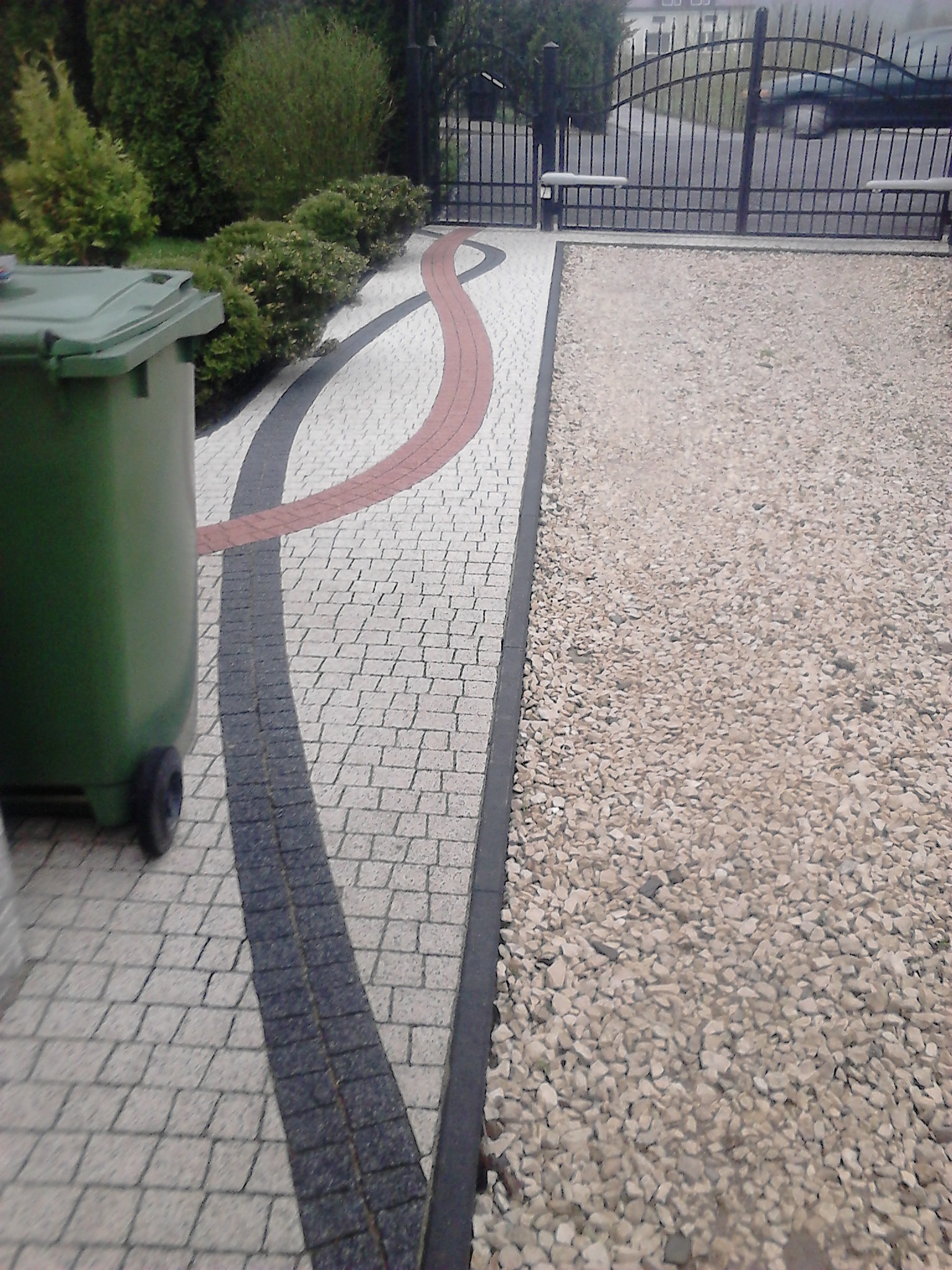 Ułożona kostka brukowa w odcieniach szarości, czerni i czerwieni tworząca ścieżkę z dekoracyjnym wzorem falistym, obok zielony pojemnik na śmieci na kółkach i fragment drogi wysypanej żwirem...