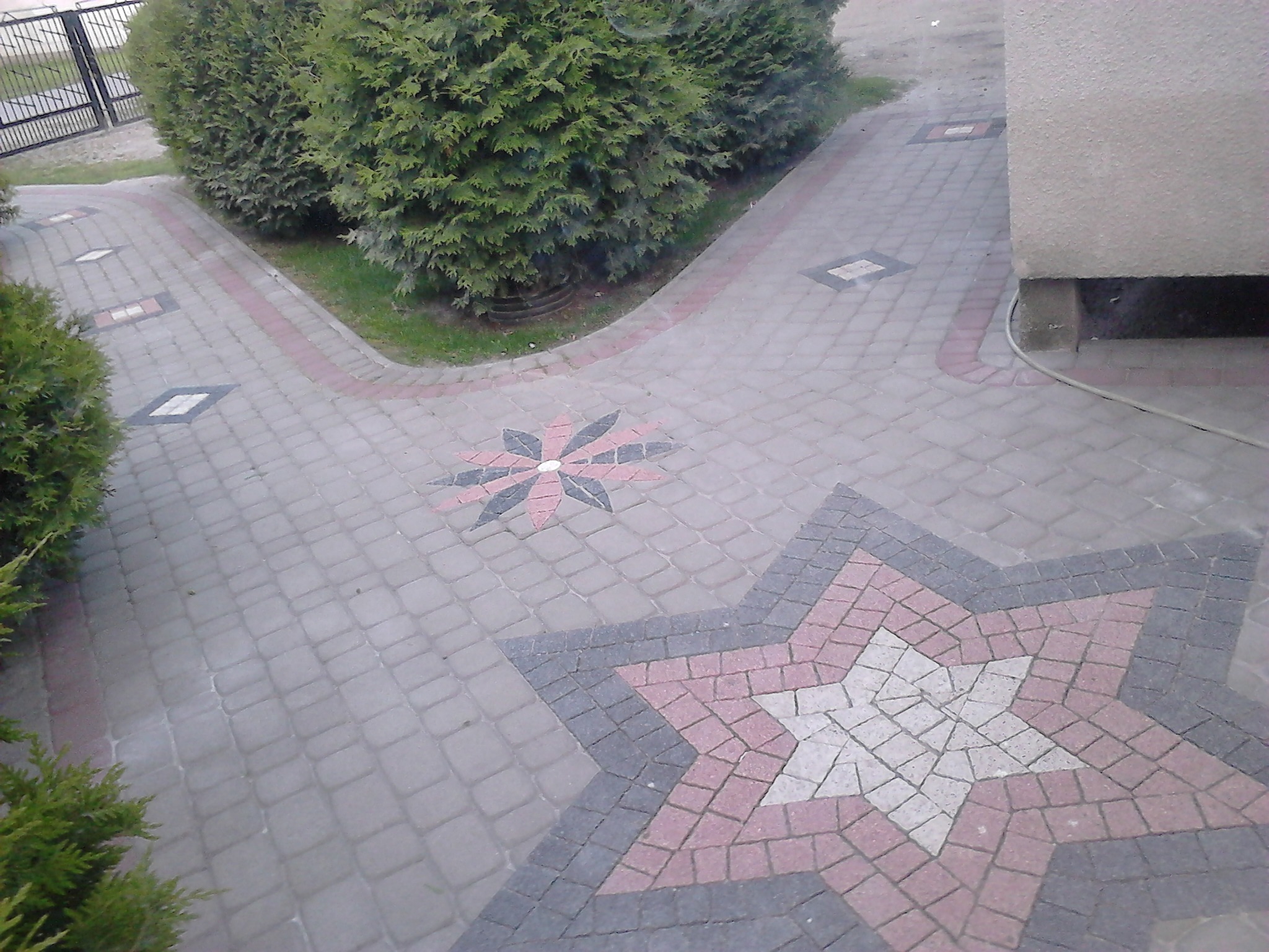 Ułożona ścieżka z kostki brukowej w kształcie rozchodzących się dróg, z dekoracyjnymi wzorami gwiazd i kwiatów z kostki w kolorach szarym, różowym i białym, obrzeża z czerwonej kostki, otoczone...
