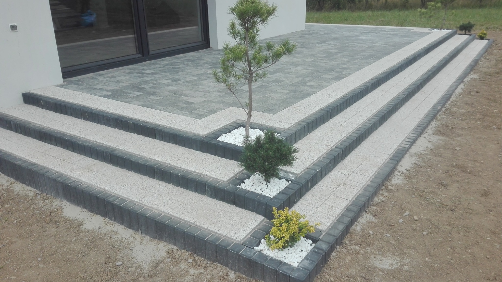 Schody wejściowe do domu wyłożone jasnoszarą kostką brukową z ciemnoszarą obwódką, z trzema miniaturowymi drzewkami iglastymi i krzewami ozdobnymi w narożach, wysypane białym kamieniem dekoracyjnym.
