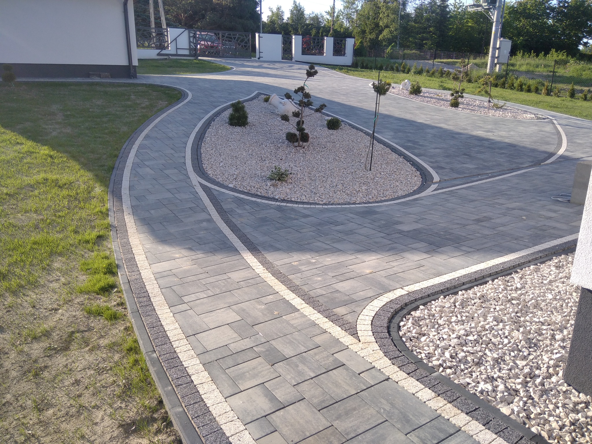 Ułożona z szarej kostki brukowej, kręta ścieżka z jasnymi obrzeżami, otaczająca rabatę z białym żwirem i ozdobnymi drzewkami, prowadząca do domu z bramą w tle.