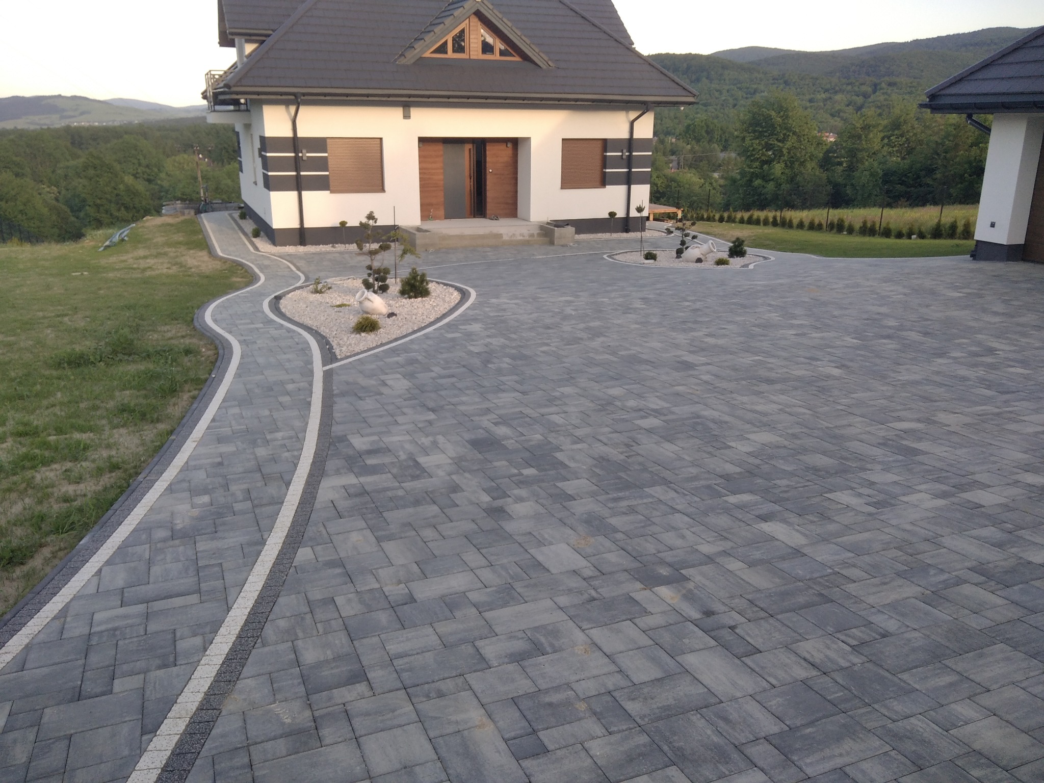 Szara kostka brukowa ułożona na podjeździe do nowoczesnego domu z ciemnym dachem, widoczne dekoracyjne obrzeża z jasnej kostki i ozdobne nasadzenia w żwirze, w tle zalesione wzgórza.