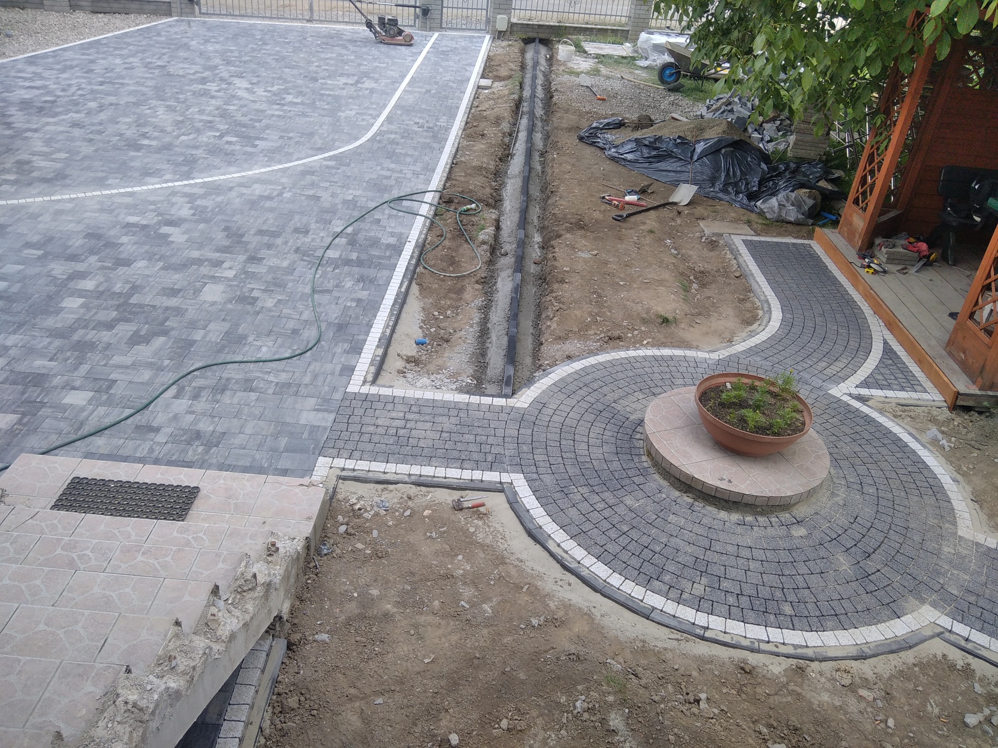 Widok z góry na fragment posesji w trakcie prac brukarskich: ułożona szara kostka na placu i ścieżce z dekoracyjnym okręgiem z donicą pośrodku, wykopany rów drenażowy wzdłuż placu, widoczna...