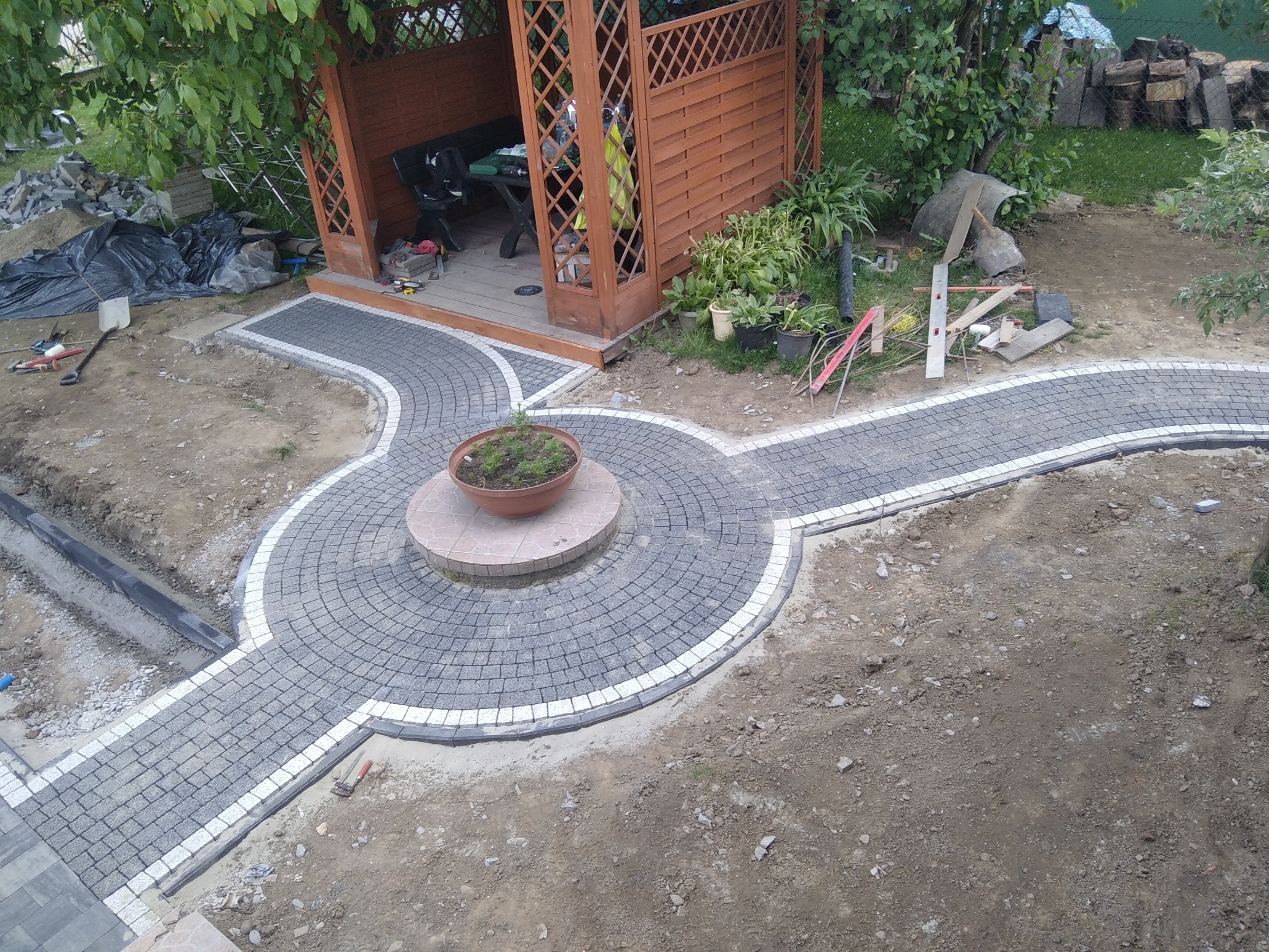 Widok z góry na ścieżkę z kostki brukowej w ogrodzie, z okrągłym placem pośrodku, na którym stoi donica z rośliną; w tle widoczna drewniana altana i częściowo ułożona kostka z widocznym drenażem.