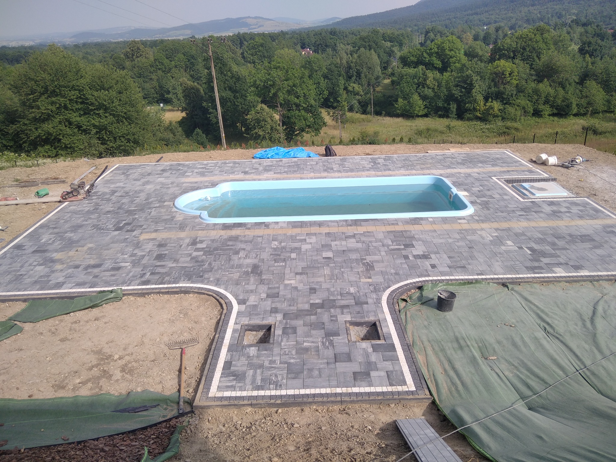 Ułożona kostka brukowa wokół basenu ogrodowego w trakcie prac wykończeniowych, widoczne narzędzia, teren częściowo zabezpieczony plandeką, w tle zalesione wzgórza.