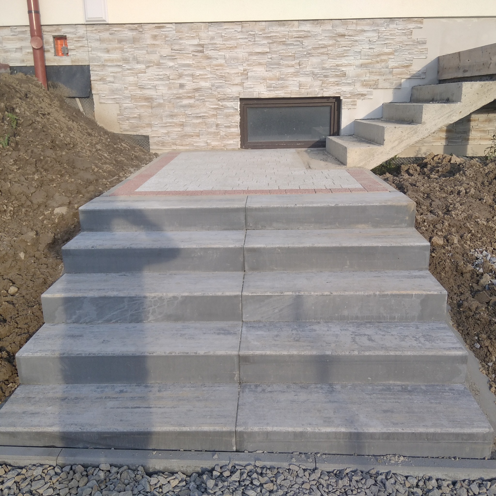 Schody z szarych betonowych płyt prowadzące do budynku z elewacją z kamienia dekoracyjnego, widoczny fragment okna piwnicznego i nasyp ziemi po bokach.