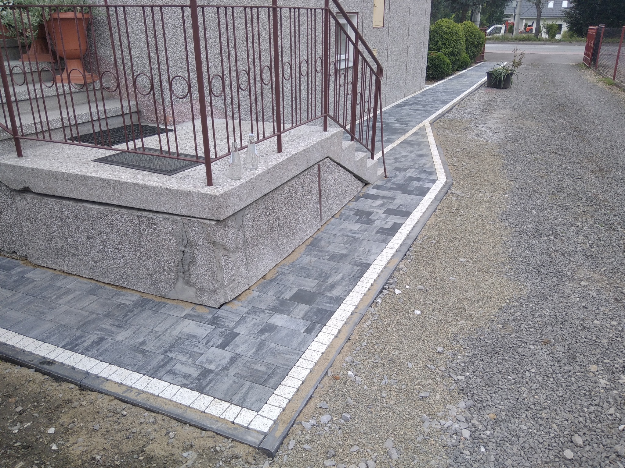 Ułożona szara kostka brukowa z białym obramowaniem prowadząca od schodów wejściowych do domu, obok utwardzona droga z kruszywa, dwie szklane butelki na schodach.