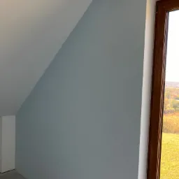 Ściana w odcieniach szarości w pokoju na poddaszu po malowaniu, widoczne gniazdka elektryczne i drewniana rama okienna z widokiem na zewnątrz.