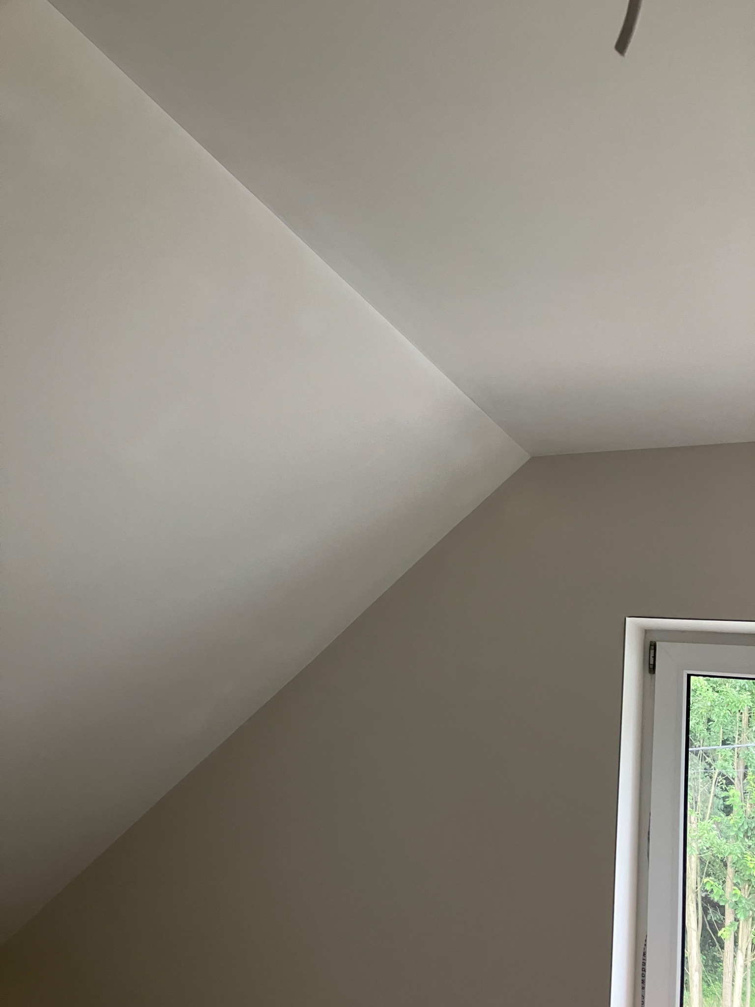 Poddasze po remoncie, ściana pomalowana na szaro, sufit na biało, widoczne okno z widokiem na drzewa.