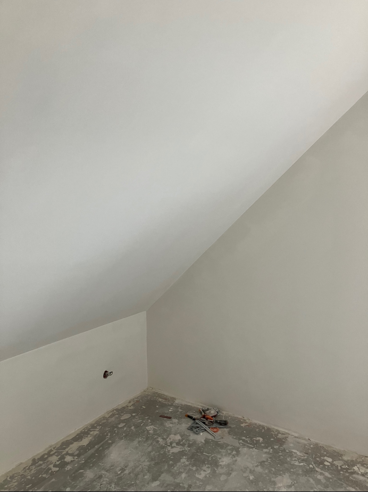 Poddasze po szpachlowaniu i wstępnym malowaniu, widoczne ślady na podłodze i narzędzia przy ścianie, przygotowane gniazdko elektryczne.