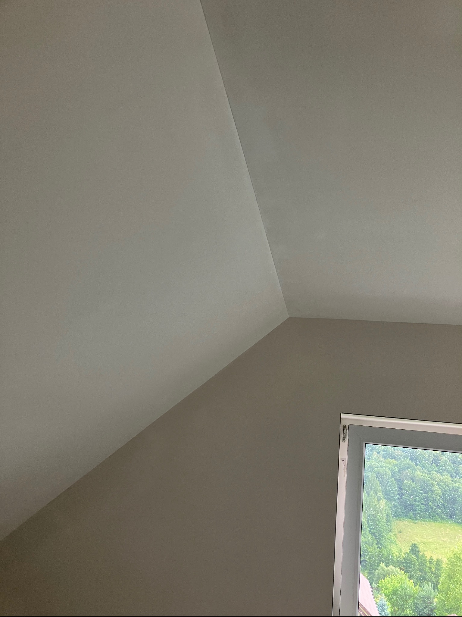 Pomalowane na szaro ściany i biały sufit na poddaszu ze skosem, widok na zewnątrz przez okno.