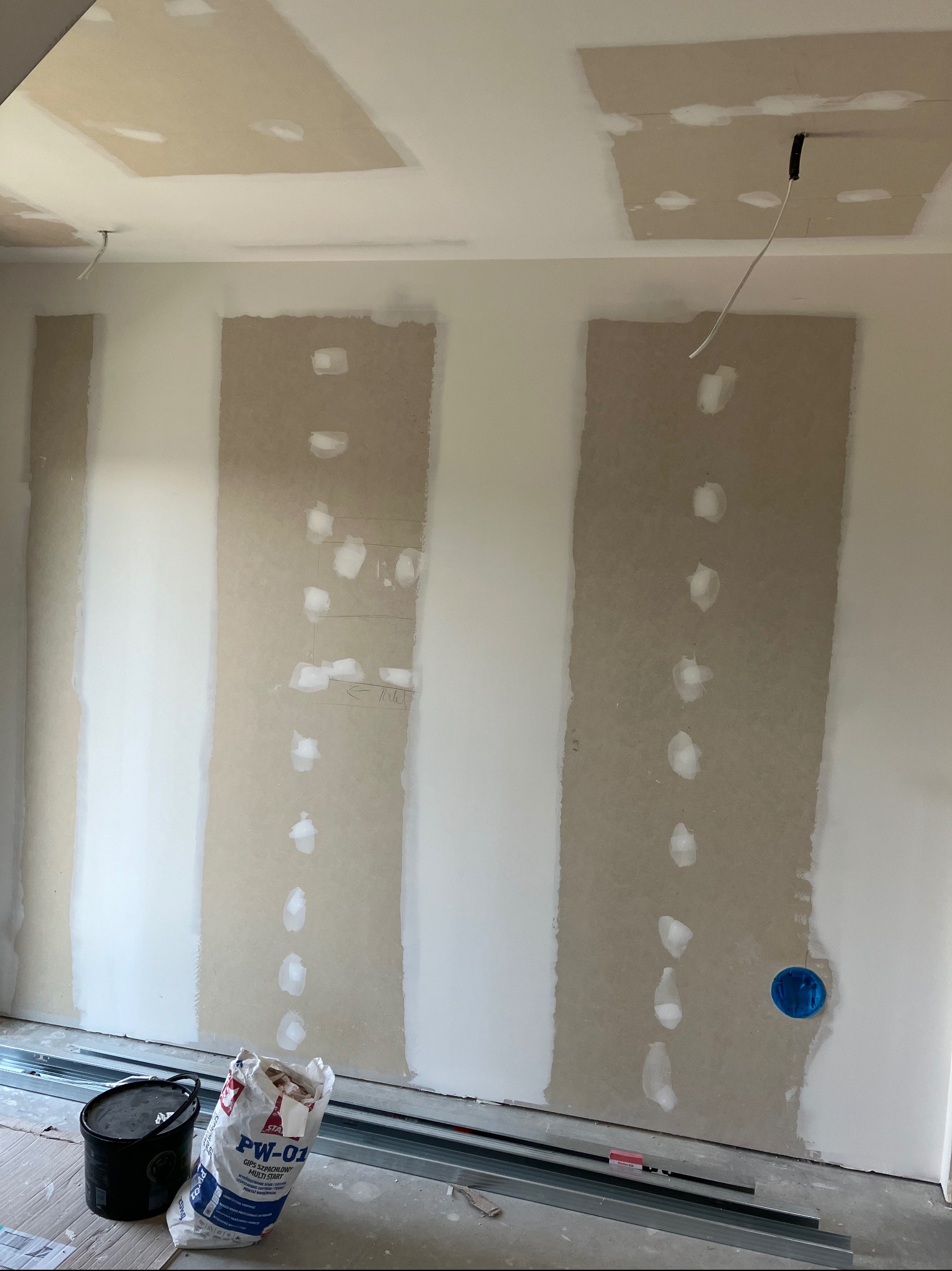 Wnętrze w trakcie prac wykończeniowych: płyty gipsowo-kartonowe ze śladami szpachlowania, widoczne puszki elektryczne, profile metalowe, wiadro i worek z gipsem szpachlowym PW-01.
