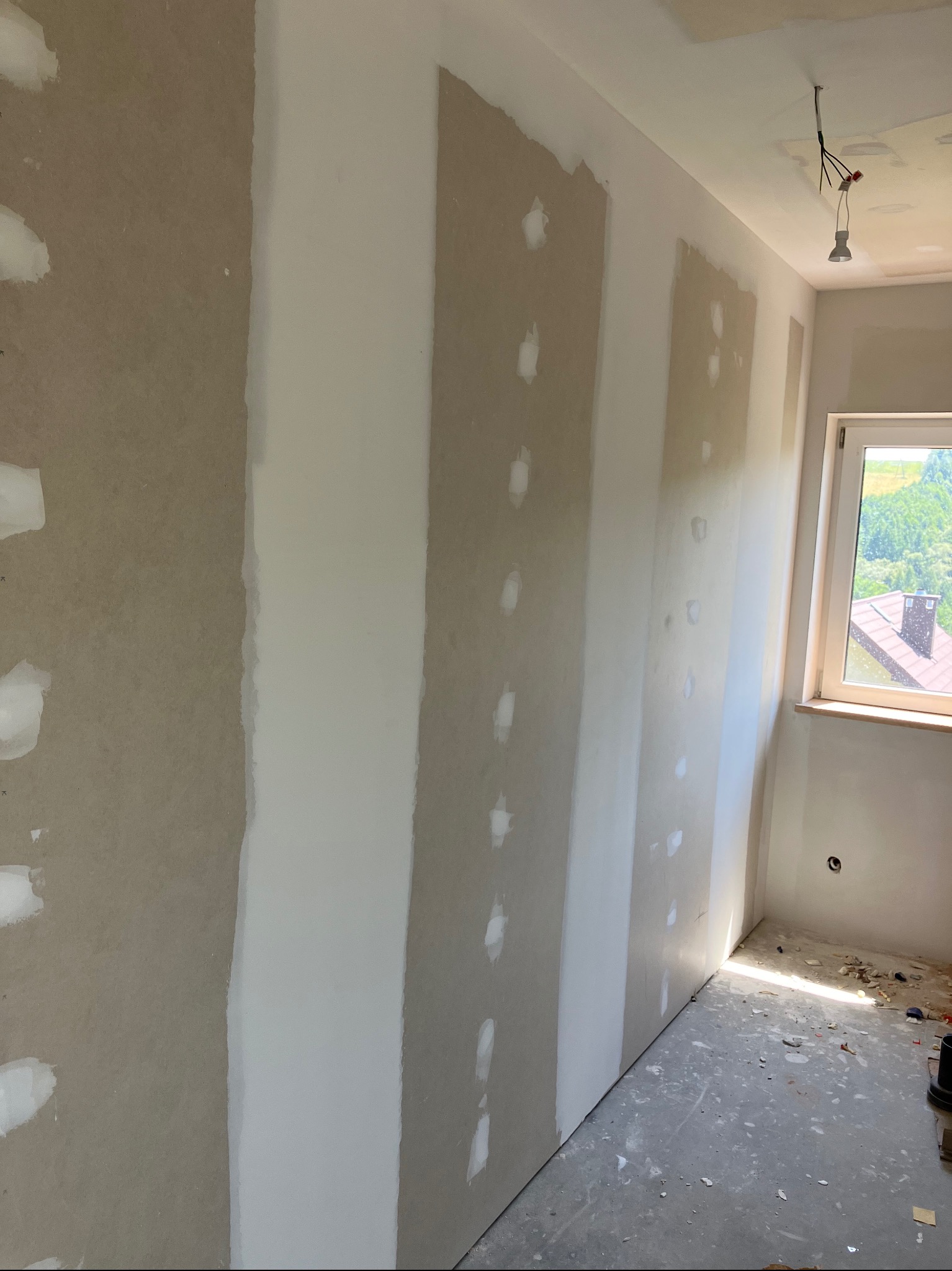 Ściana z płyt gipsowo-kartonowych w trakcie szpachlowania, widoczne miejsca z nałożoną gładzią, surowy beton na podłodze, okno z widokiem na zieleń.