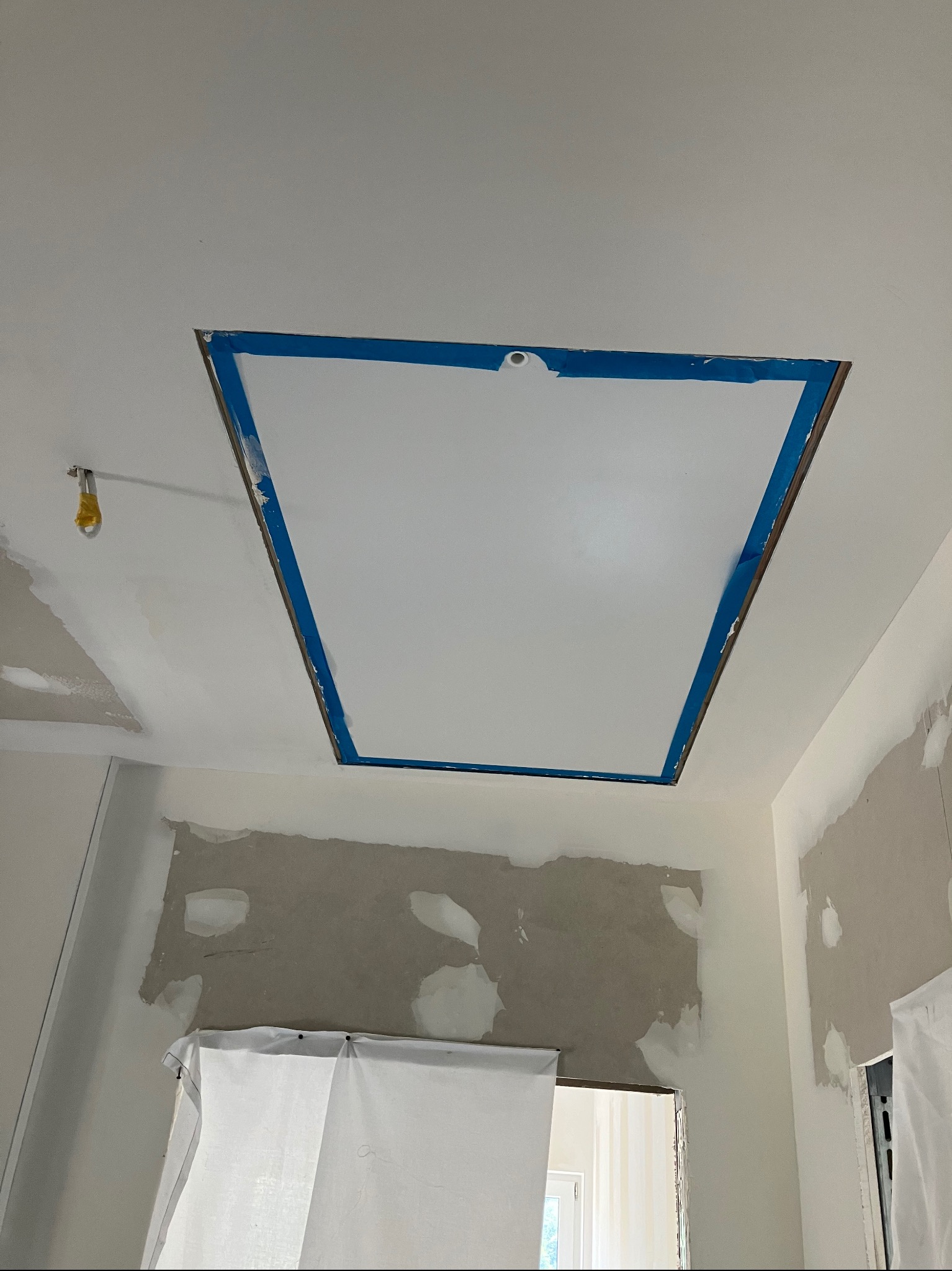 Przygotowanie sufitu do malowania: wycięty otwór zabezpieczony niebieską taśmą malarską, widoczne ślady szpachlowania na ścianach i zakryte folią drzwi.
