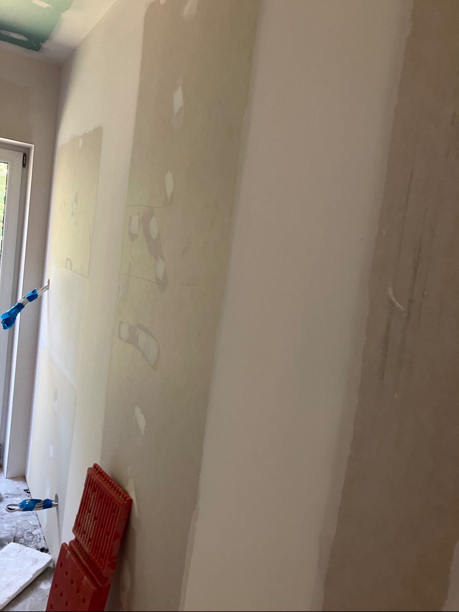 Ściana po szpachlowaniu, z widocznymi łatami i przygotowaniem do malowania, zabezpieczone kable elektryczne taśmą, w tle widoczne okno.