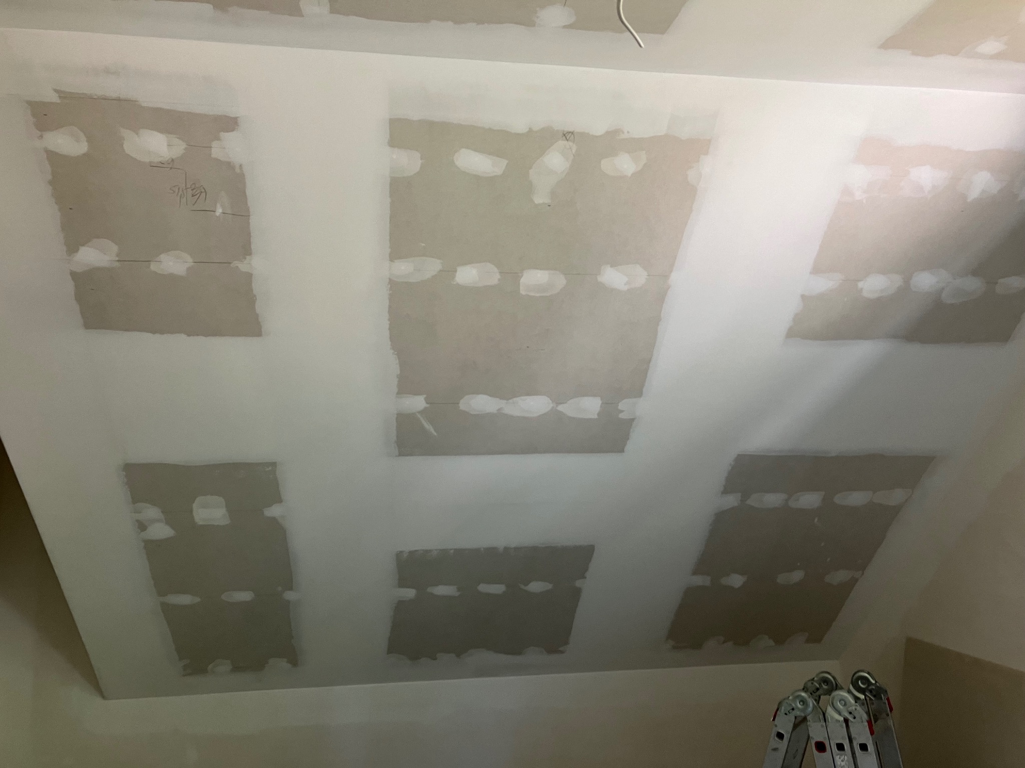 Sufit poddasza z widocznymi łatami płyt kartonowo-gipsowych, częściowo pokrytymi masą szpachlową, przygotowany do dalszego wykończenia, z fragmentem drabiny w dolnym rogu.