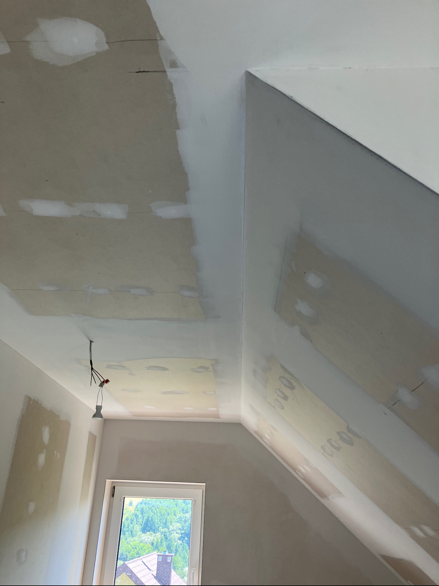 Poddasze w trakcie remontu: ściany i skośny sufit z widocznymi łatami szpachli, przygotowane do dalszych prac wykończeniowych, z oknem w tle.