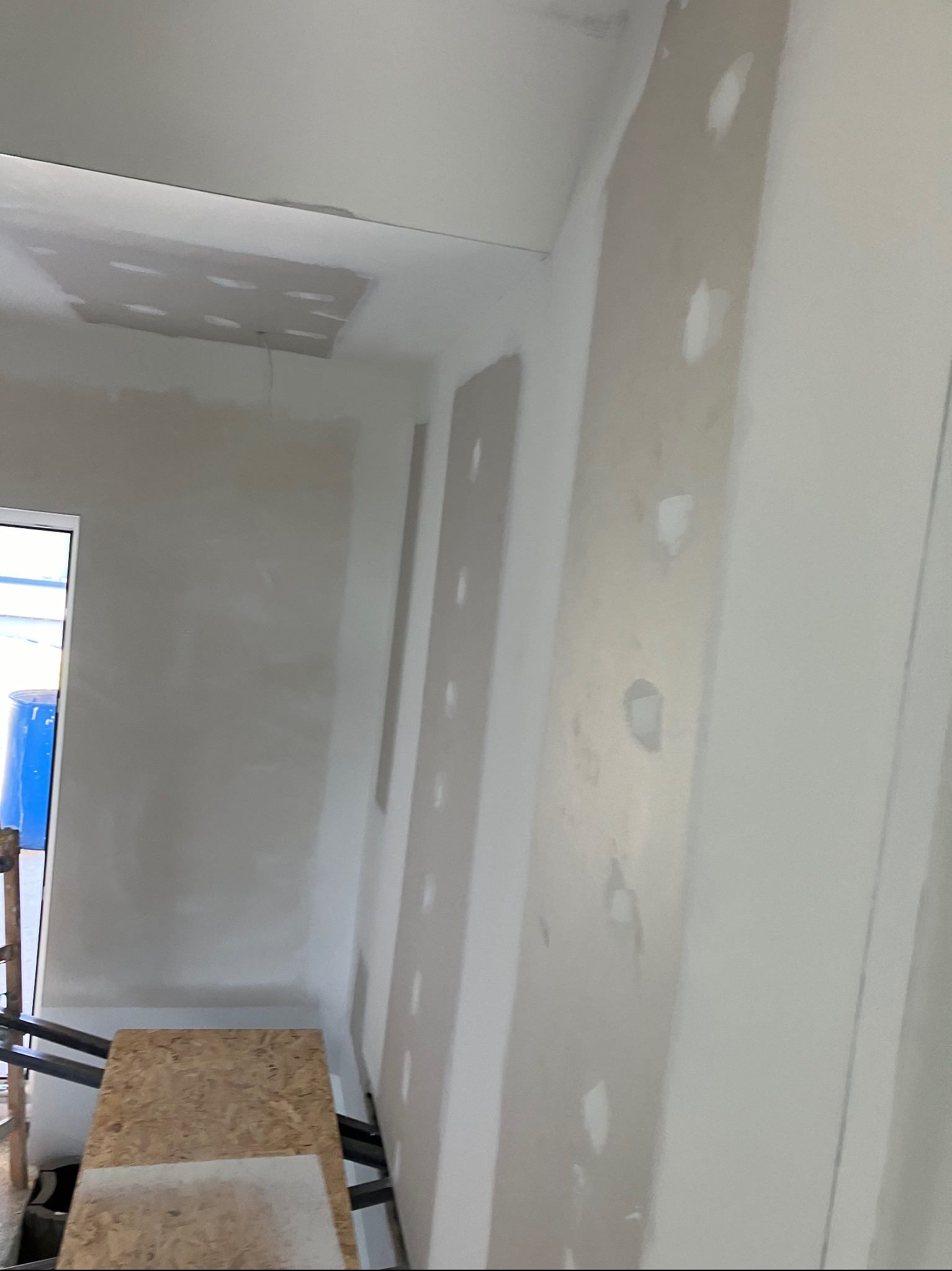 Ściana po szpachlowaniu z widocznymi punktowymi wypełnieniami ubytków, przygotowana do dalszych prac wykończeniowych, widoczny fragment rusztowania z deską.