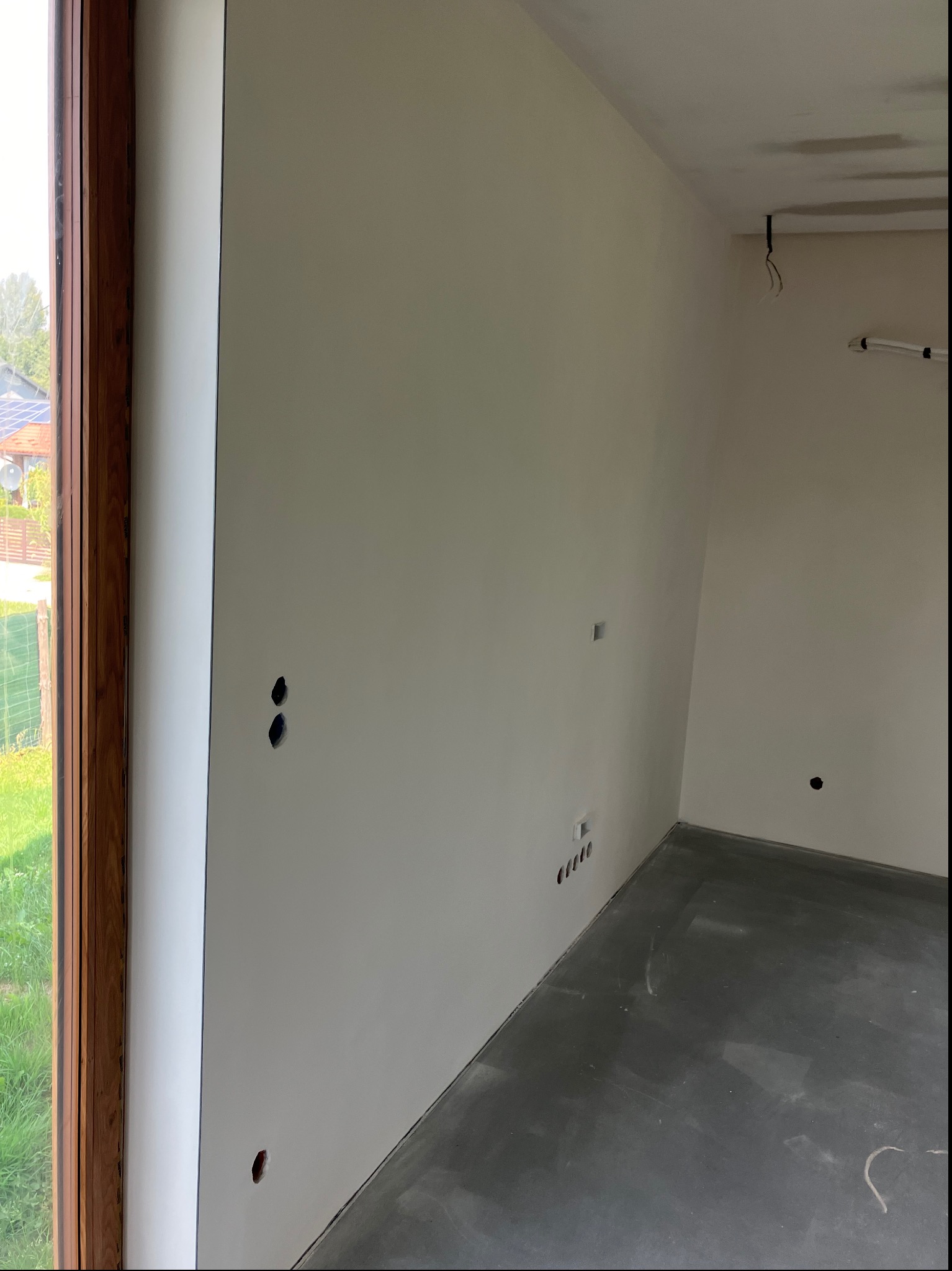 Wnętrze w stanie deweloperskim z gładkimi, jasnymi ścianami, surową wylewką na podłodze, widocznymi otworami na gniazdka elektryczne i fragmentem drewnianego okna z widokiem na zieleń.