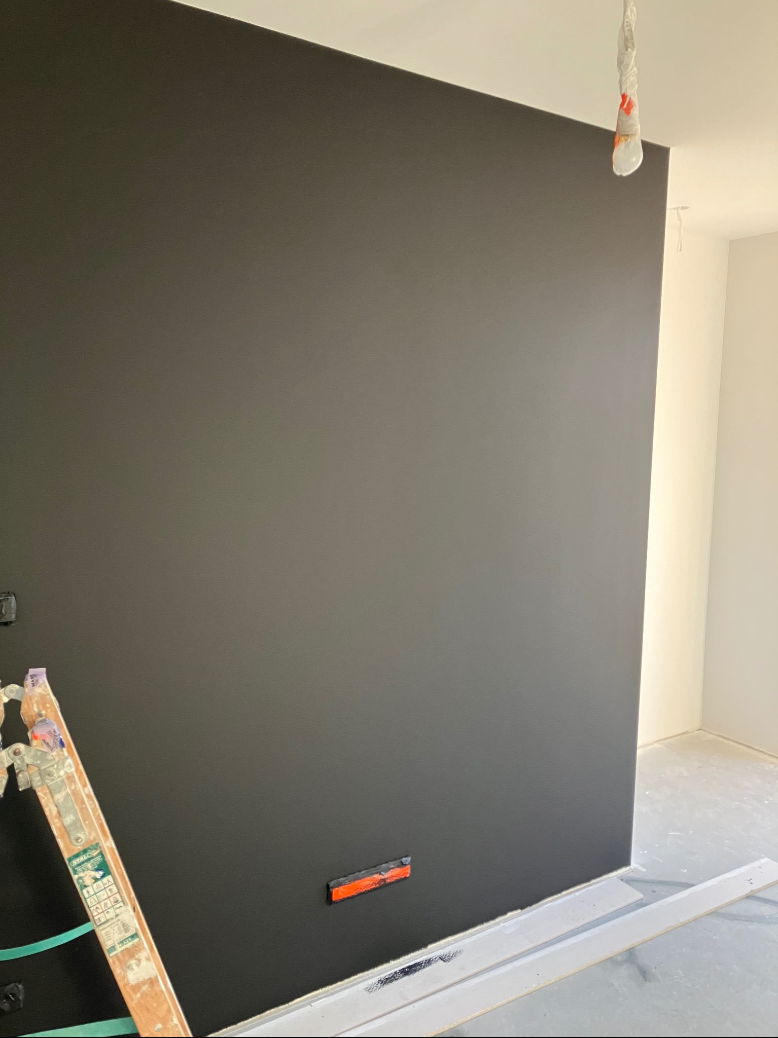 Świeżo pomalowana, gładka, czarna ściana w pokoju z białą listwą przypodłogową i drabiną malarską w rogu, widoczny fragment instalacji elektrycznej.