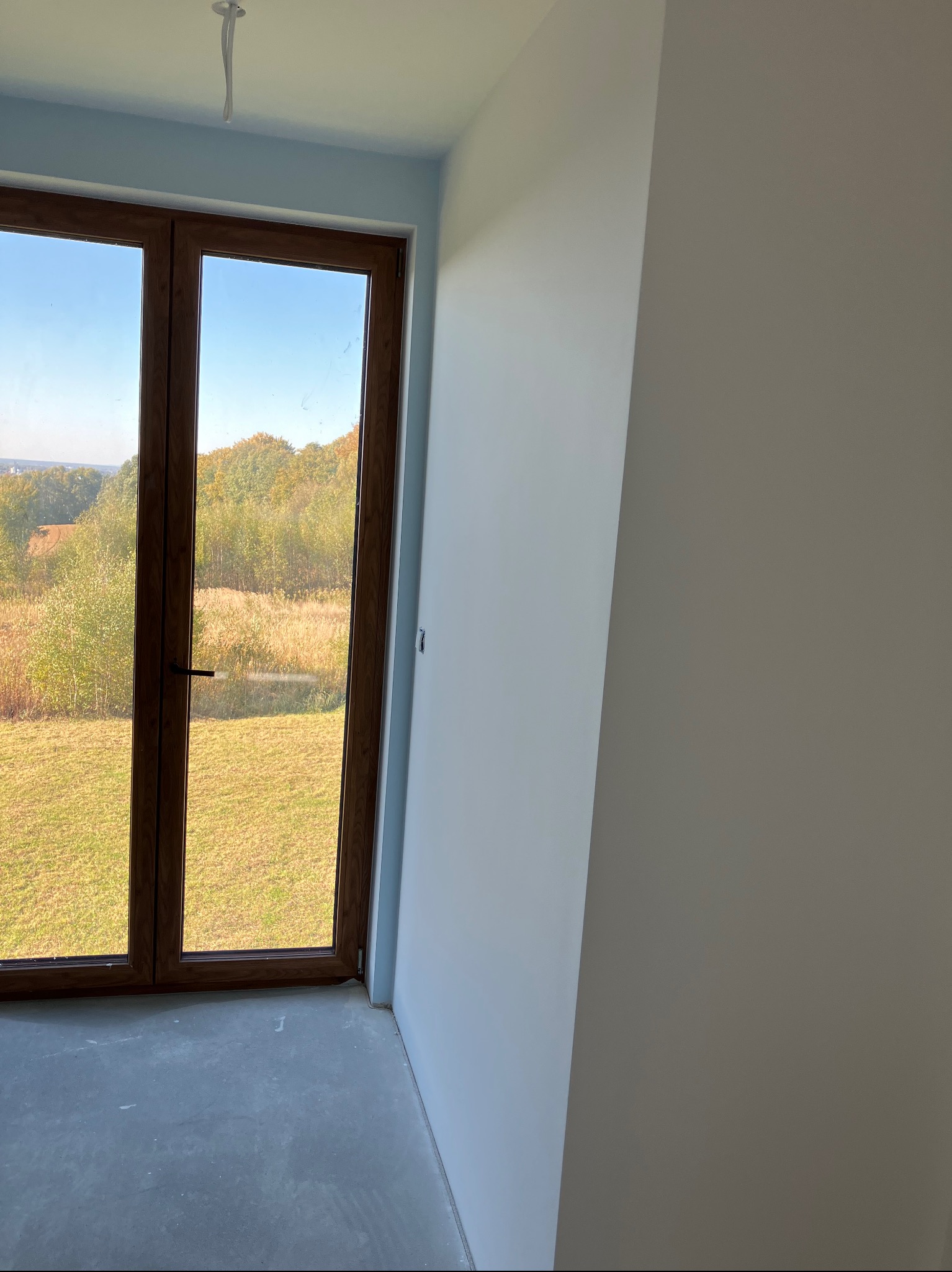 Wnętrze pokoju z widokiem na zewnątrz przez okno balkonowe, ściany pomalowane na biało, sufit z widocznym kablem oświetleniowym, podłoga betonowa.