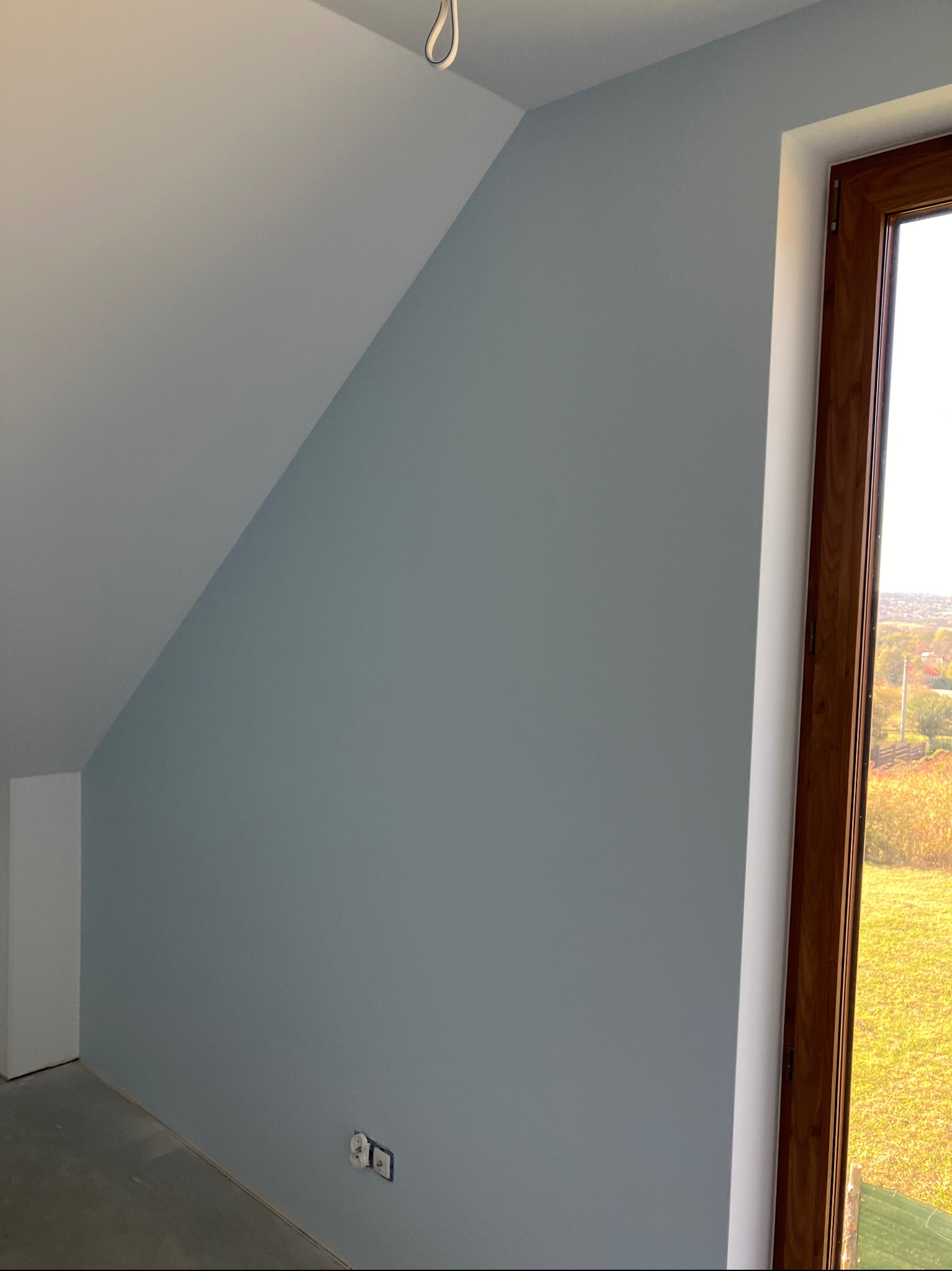 Ściana w odcieniach szarości w pokoju na poddaszu po malowaniu, widoczne gniazdka elektryczne i drewniana rama okienna z widokiem na zewnątrz.