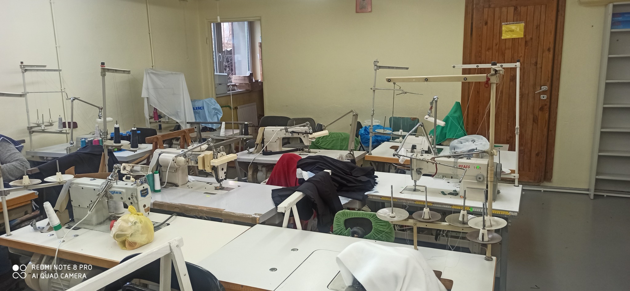 Wnętrze szwalni z kilkoma stanowiskami pracy wyposażonymi w maszyny do szycia, widoczne materiały i nici, w tle drzwi i regał.