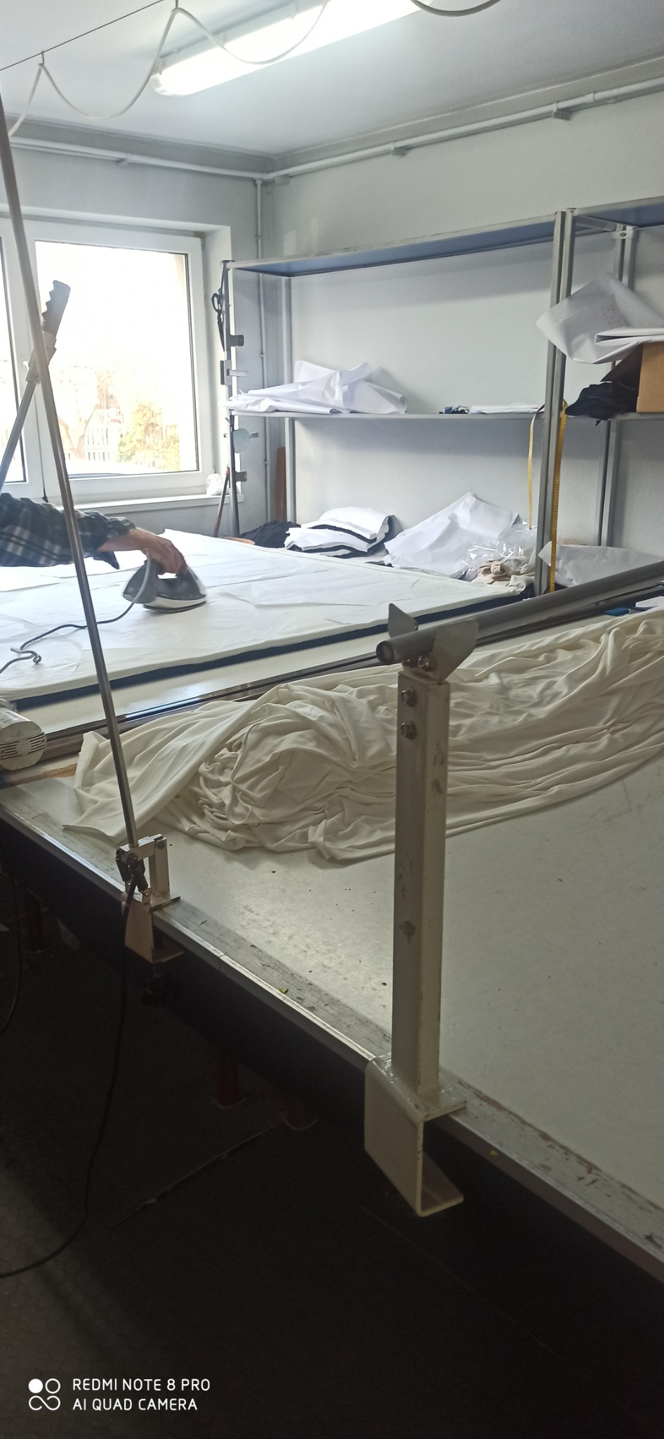 Prasowanie jasnej tkaniny na stole krojczym w pracowni, widoczne stosy wykrojonych elementów na półkach w tle.