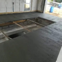 Wylewka betonowa w trakcie wykonywania, widoczne drewniane elementy szalunku i instalacje w ścianie budynku w stanie surowym.