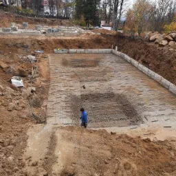 Przygotowanie zbrojenia na dużej płycie fundamentowej, widoczny pracownik w niebieskiej kurtce, teren budowy z wykopem i elementami szalunku.