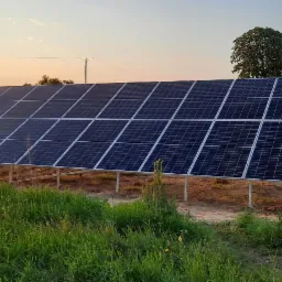 Instalacja fotowoltaiczna na metalowych podporach, umieszczona na łące z wysoką trawą, w tle drzewa i zachodzące słońce.