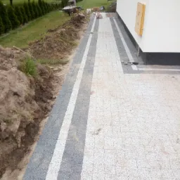 Świeżo ułożona, jasna kostka brukowa z ciemnoszarym obramowaniem i pasami, przylegająca do białej ściany budynku, widoczna kupa ziemi i taczka w tle.