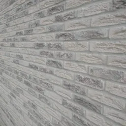 Ściana z jasnoszarej cegły klinkierowej z wyraźną fakturą, ułożona w regularny wzór, widziana pod kątem z widocznym fragmentem roślinności u dołu.