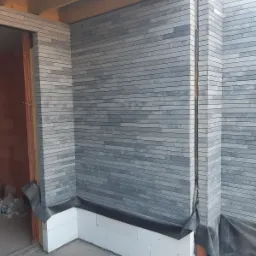 Montaż elewacji z szarej, wąskiej cegły klinkierowej na narożniku budynku, widoczne białe bloczki fundamentowe i czarna folia izolacyjna.